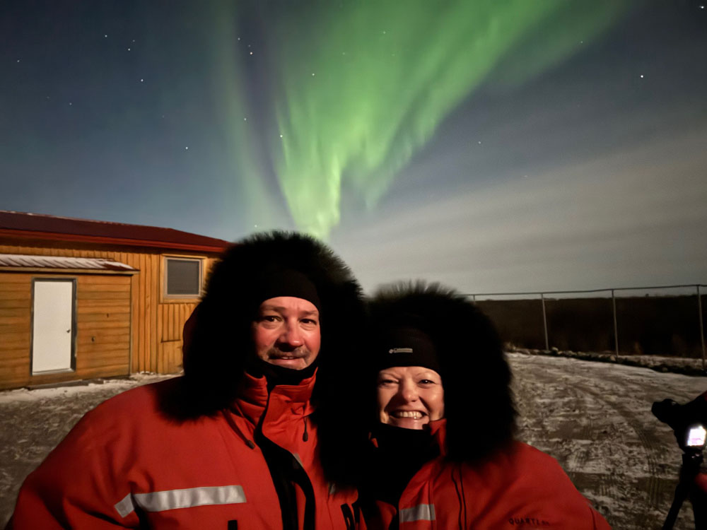 Dave Bouskill and Deb Corbeil of The Planet D in front of northern lights at Seal River Heritage Lodge.