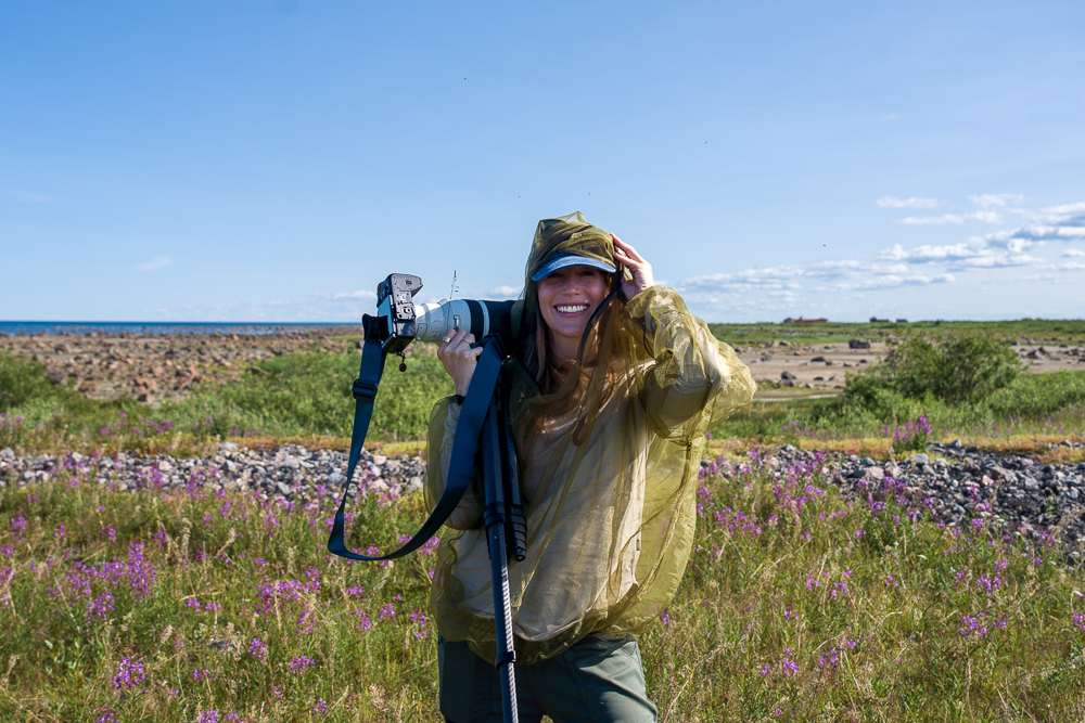 Erin Sobon with camera on the rocky coast at Seal River Heritage Lodge with a backdrop of fireweed.